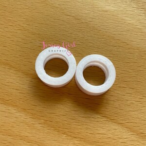 May include: Two white, circular objects with a hole in the center, resembling small rings. The objects are placed on a light brown wooden surface. The text "Jersey Girl Graphics" is visible in the image.