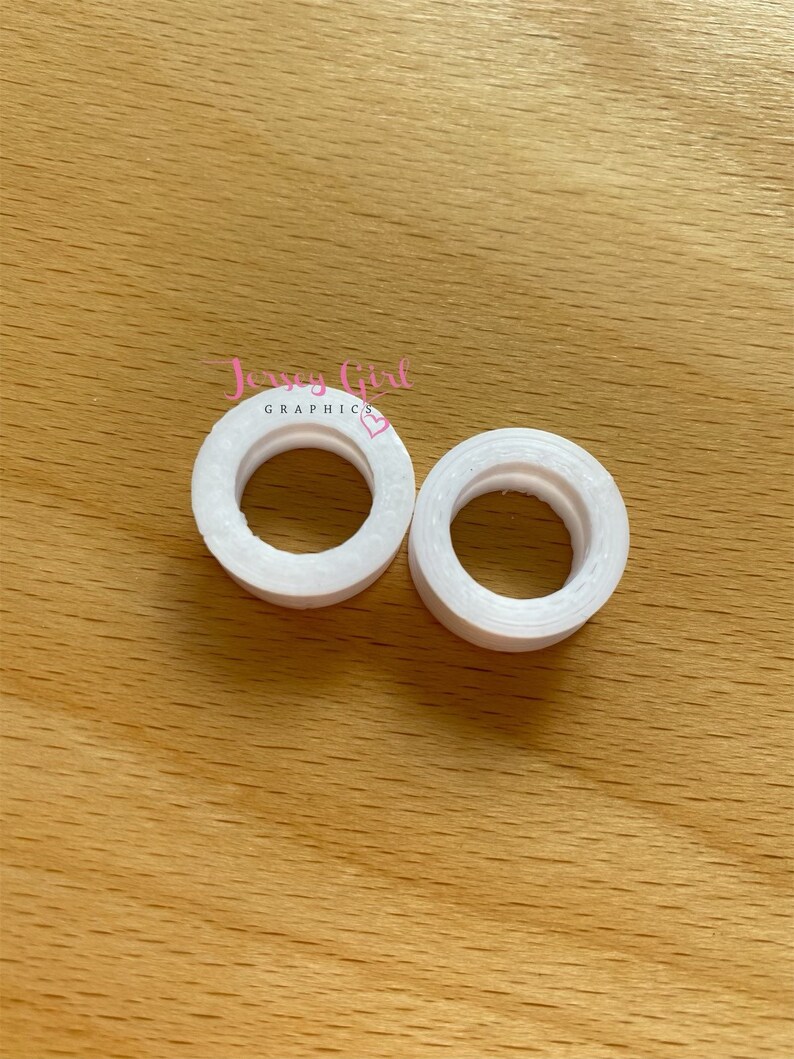 May include: Two white, circular objects with a hole in the center, resembling small rings. The objects are placed on a light brown wooden surface. The text "Jersey Girl Graphics" is visible in the image.