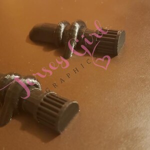 Op de afbeelding: Twee zwarte wijnflesstoppers met een uniek ontwerp. De stoppers hebben een getextureerd, cilindrisch lichaam met een geribbelde uiteinde en een decoratief knoopdetail. De achtergrond is effen bruin.