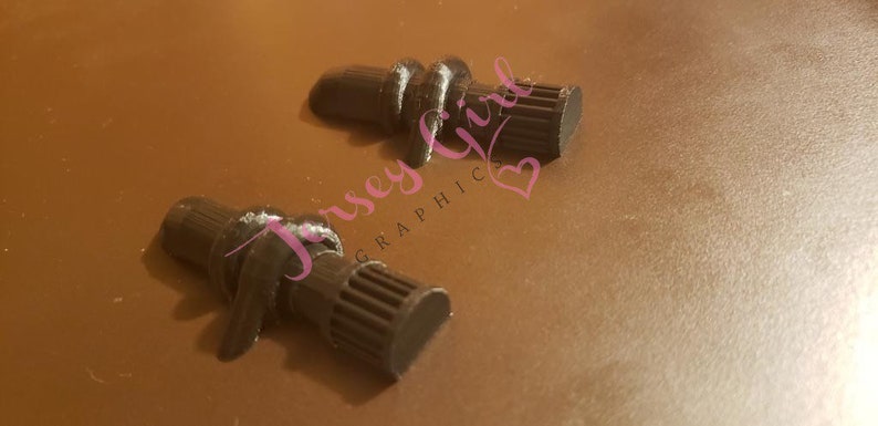 Op de afbeelding: Twee zwarte wijnflesstoppers met een uniek ontwerp. De stoppers hebben een getextureerd, cilindrisch lichaam met een geribbelde uiteinde en een decoratief knoopdetail. De achtergrond is effen bruin.