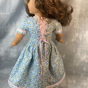 Puede incluir: Un vestido floral azul y blanco con una cinta rosa y ribete de encaje. El vestido tiene una espalda con cordones y est&aacute; dise&ntilde;ado para una mu&ntilde;eca.