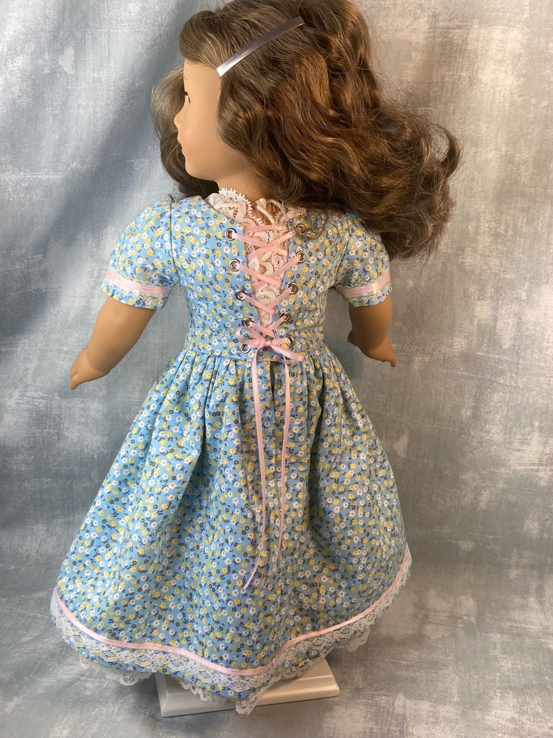 Puede incluir: Un vestido floral azul y blanco con una cinta rosa y ribete de encaje. El vestido tiene una espalda con cordones y est&aacute; dise&ntilde;ado para una mu&ntilde;eca.