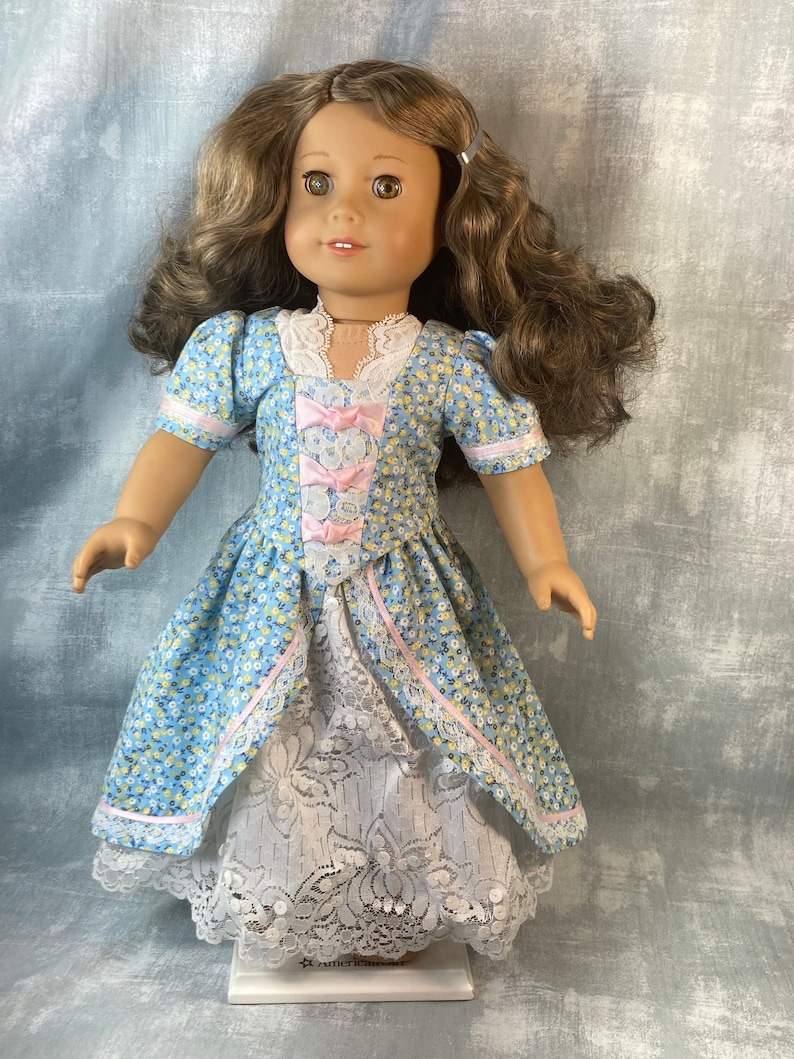 Puede incluir: Una mu&ntilde;eca con un vestido floral azul y blanco con detalles rosas y una falda de encaje blanca. La mu&ntilde;eca tiene el pelo casta&ntilde;o y los ojos marrones. La mu&ntilde;eca est&aacute; de pie sobre una base blanca con el texto "American Girl" impreso en ella.