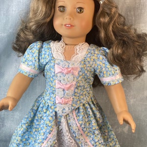 Puede incluir: Una mu&ntilde;eca con un vestido floral azul y blanco con detalles de cinta rosa y un borde de encaje blanco. La mu&ntilde;eca tiene cabello casta&ntilde;o y ojos marrones.