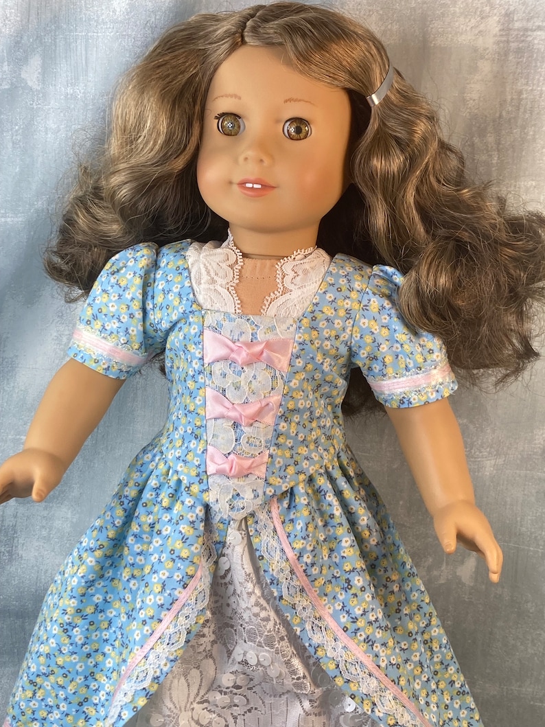 Puede incluir: Una mu&ntilde;eca con un vestido floral azul y blanco con detalles de cinta rosa y un borde de encaje blanco. La mu&ntilde;eca tiene cabello casta&ntilde;o y ojos marrones.