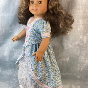 Puede incluir: Una mu&ntilde;eca con un vestido floral azul y blanco con una cinta rosa y ribete de encaje. El vestido tiene una enagua de encaje blanco. La mu&ntilde;eca tiene el pelo casta&ntilde;o y los ojos marrones.