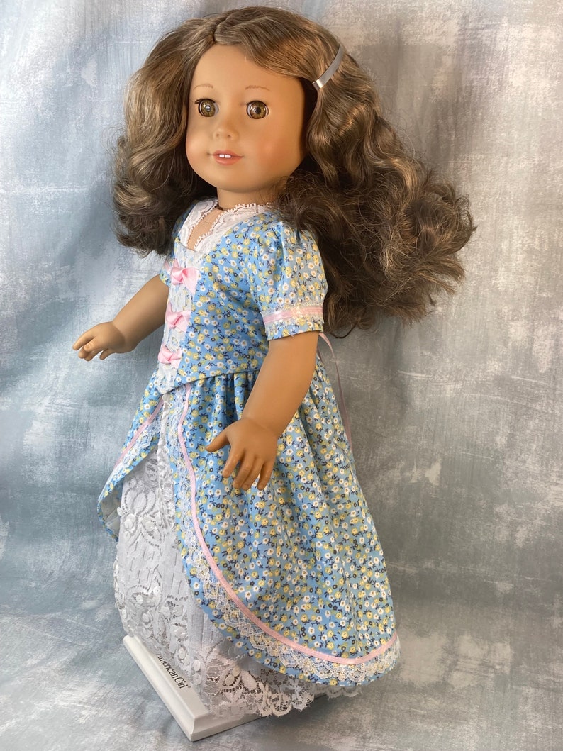 Puede incluir: Una mu&ntilde;eca con un vestido floral azul y blanco con una cinta rosa y ribete de encaje. El vestido tiene una enagua de encaje blanco. La mu&ntilde;eca tiene el pelo casta&ntilde;o y los ojos marrones.