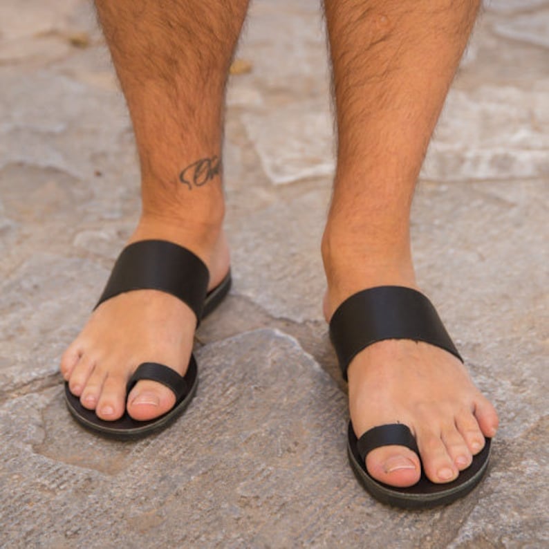 May include: Black leather sandals with a toe loop and wide strap across the top of the foot. The sandals are worn on a stone surface.