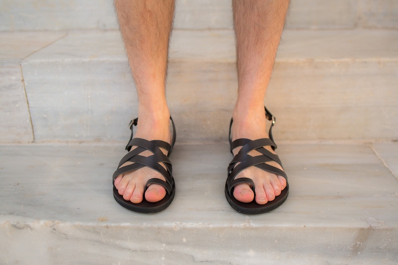 May include: Black leather sandals with a crisscross design over the foot and a toe loop. The sandals have a buckle closure and are worn on a marble surface. The sandals are a dark brown color.