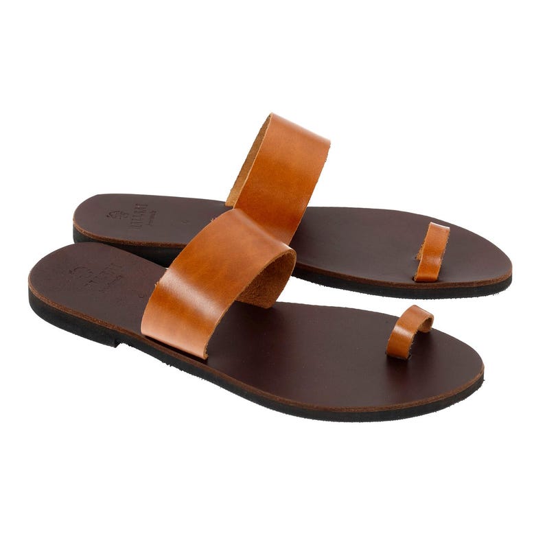 May include: A pair of brown leather sandals with a toe loop and a wide strap across the top of the foot. The sandals have a dark brown sole and a light brown leather upper. The sandals are designed for comfort and style.