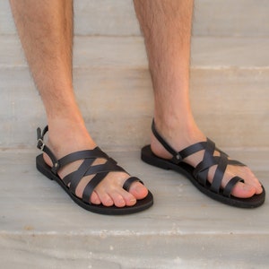 May include: Black leather sandals with a strappy design. The sandals have a toe loop and an adjustable ankle strap with a buckle. The soles are flat and black. The sandals are shown on a light-colored stone surface.