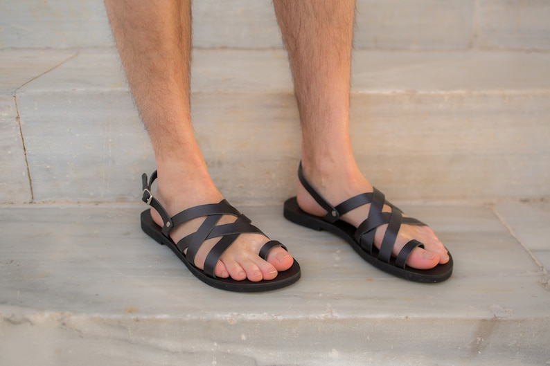 May include: Black leather sandals with a strappy design. The sandals have a toe loop and an adjustable ankle strap with a buckle. The soles are flat and black. The sandals are shown on a light-colored stone surface.