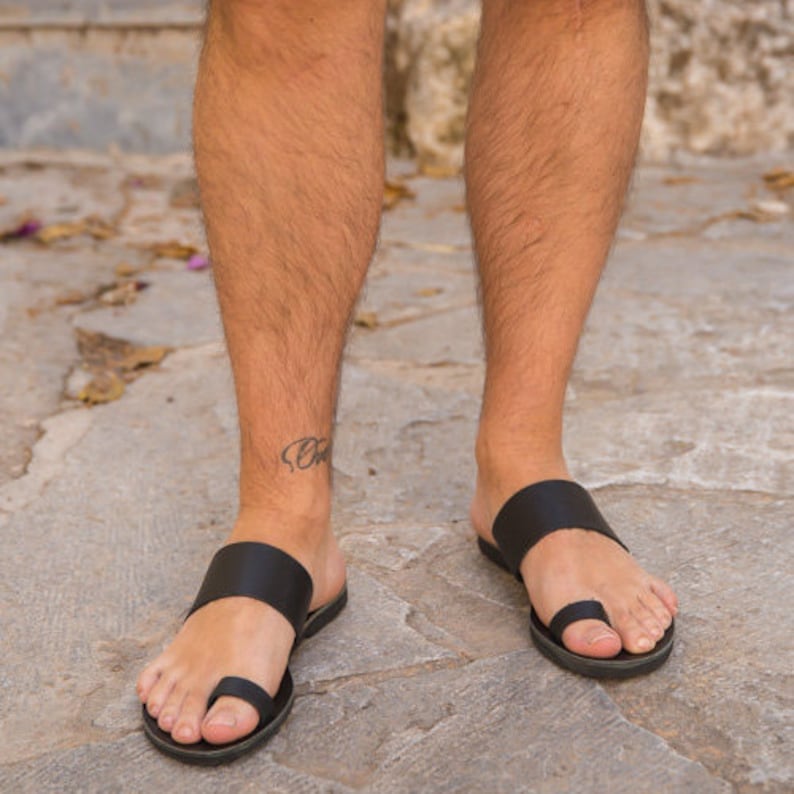 May include: Black leather sandals with a toe loop and wide strap across the top of the foot. The sandals are worn on a stone surface.