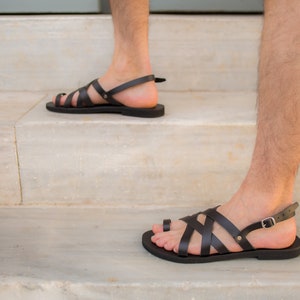May include: Black leather sandals with multiple straps and a toe loop. The sandals have a buckle closure and are worn on a person's feet. The sandals are on a white marble staircase.