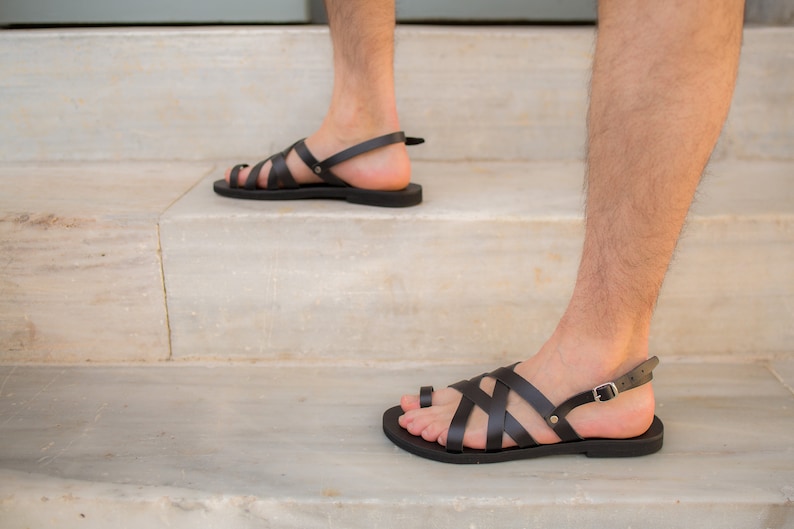 May include: Black leather sandals with multiple straps and a toe loop. The sandals have a buckle closure and are worn on a person's feet. The sandals are on a white marble staircase.