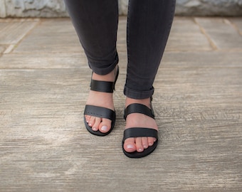 May include: A pair of black leather sandals with two straps across the foot. The sandals have a simple design and are worn on a wooden surface.