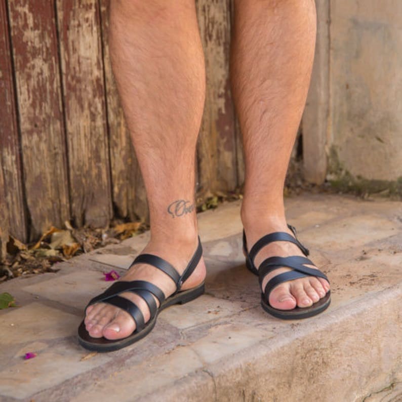 Op de afbeelding: Een paar zwarte leren sandalen met gekruiste banden. De sandalen worden gedragen aan de voeten van een persoon.