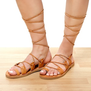 May include: Tan leather sandals with multiple straps and ankle-high wrap-around ties. The sandals have a flat sole and are displayed on a light wood surface. The design is reminiscent of ancient Greek footwear.