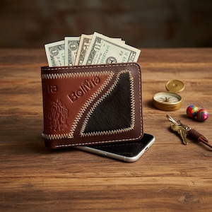 May include: Brown leather wallet with the word "Bolivia" and a stitched design. The wallet is open, revealing several U.S. dollar bills. A black smartphone is beneath the wallet. A small compass, keys, and a colorful maraca are also visible.