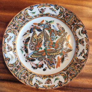 Puede incluir: Un plato de cerámica decorativo con un diseño detallado de mariposas. El plato tiene un centro blanco con un grupo de mariposas en tonos naranja, azul y amarillo. El borde está adornado con un patrón repetido de mariposas y elementos florales.