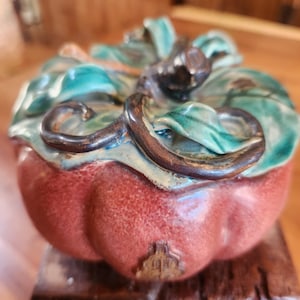 Pode incluir: Uma caixa de bugigangas de cerâmica em forma de abóbora. A caixa é principalmente vermelha com detalhes verdes e castanhos na tampa, que é projetada para se assemelhar a folhas. A caixa tem um pequeno emblema detalhado na lateral.