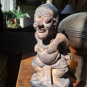 May include: A carved wooden figurine of a squatting figure with a large nose and a topknot. The figure is painted in shades of brown and black, with red accents on the eyes and mouth. The figurine is displayed on a wooden surface.