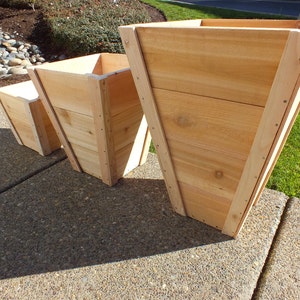 May include: Three wooden planter boxes in different sizes. The largest planter is a tall, tapered shape. The other two planters are square. All three planters are made of natural wood.
