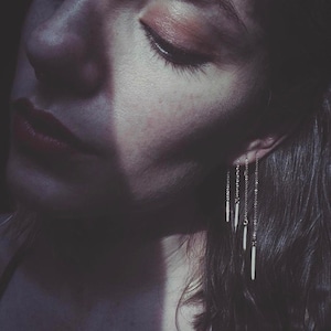 May include: A close-up of a person's face with their eye closed and a shadow cast across their cheek. They are wearing a pair of silver dangle earrings with multiple chains and small, thin, silver bars hanging from them.