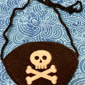 May include: A brown felt pirate eye patch with an embroidered white skull and crossbones design. The eye patch has a black elastic strap.