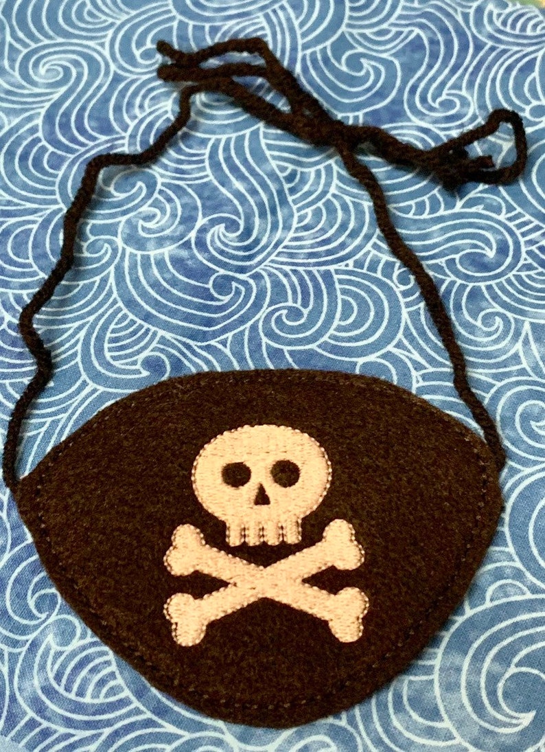 May include: A brown felt pirate eye patch with an embroidered white skull and crossbones design. The eye patch has a black elastic strap.