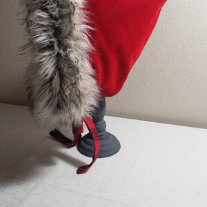 Puede incluir: Un gorro rojo con capucha y ribete de piel sint&eacute;tica. El gorro se muestra en un pedestal gris.
