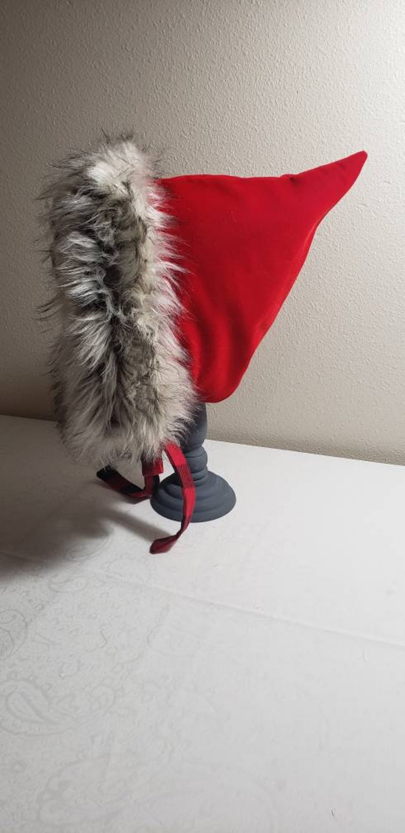 Puede incluir: Un gorro rojo con capucha y ribete de piel sint&eacute;tica. El gorro se muestra en un pedestal gris.