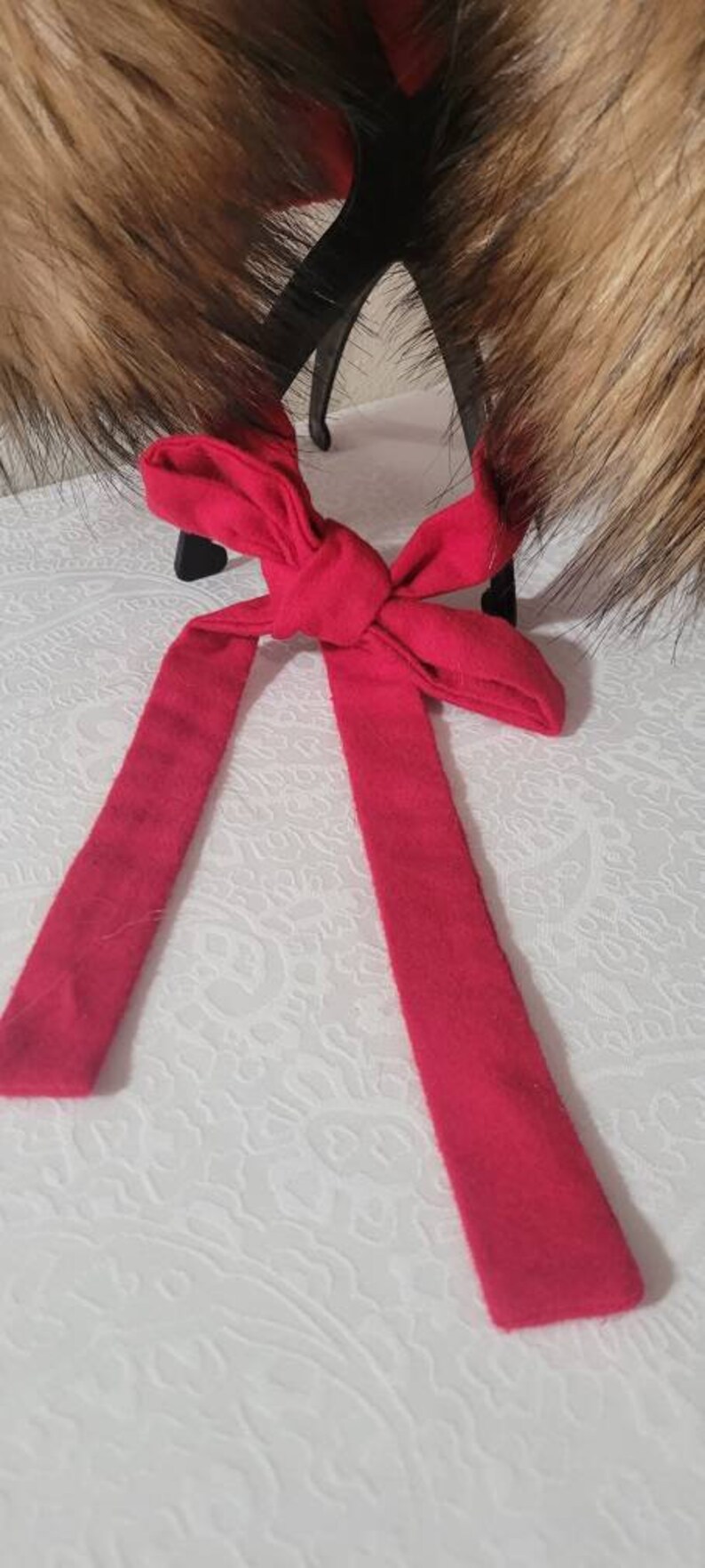 Puede incluir: Un lazo de tela rojo con un nudo en el centro. El lazo est&aacute; atado alrededor de un par de orejeras de piel marr&oacute;n.