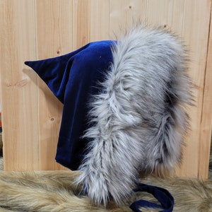 May include: A blue velvet hooded hat with a faux fur trim. The hat has a long tie that can be tied under the chin. The hat is on a brown fur surface. The tag says "Pink Owl Boutique".