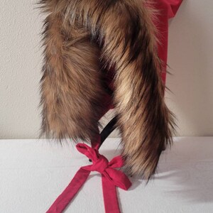 Puede incluir: Un gorro rojo con capucha y ribete de piel sint&eacute;tica marr&oacute;n. El gorro tiene una cinta roja larga atada en un lazo.