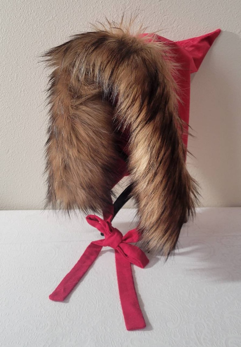 Puede incluir: Un gorro rojo con capucha y ribete de piel sint&eacute;tica marr&oacute;n. El gorro tiene una cinta roja larga atada en un lazo.