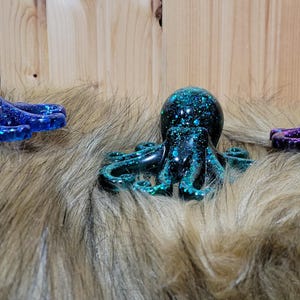 Puede incluir: Tres peque&ntilde;as figuritas de pulpo con purpurina incrustada en la resina. Los pulpos son azul, verde azulado y morado.