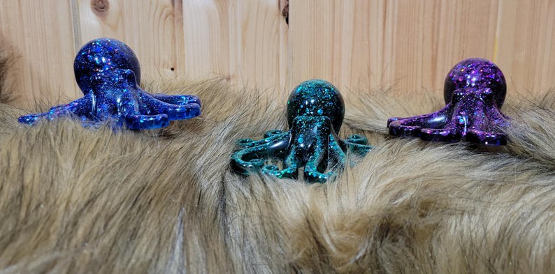 Puede incluir: Tres peque&ntilde;as figuritas de pulpo con purpurina incrustada en la resina. Los pulpos son azul, verde azulado y morado.