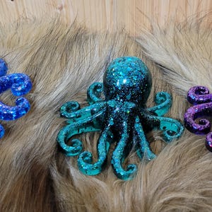 Puede incluir: Tres figuritas de pulpo con purpurina. Una es azul, otra es turquesa y otra es morada.