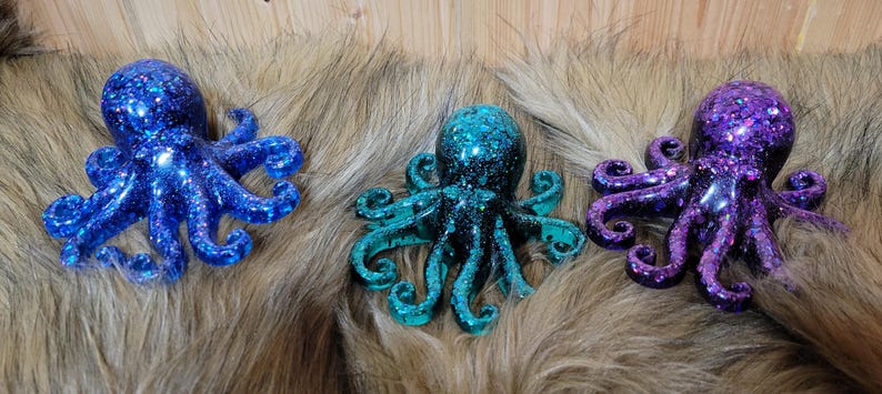 Puede incluir: Tres figuritas de pulpo con purpurina. Una es azul, otra es turquesa y otra es morada.