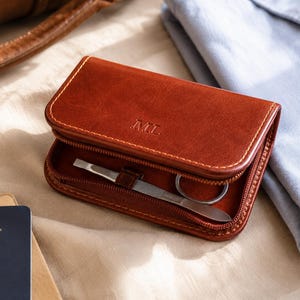 May include: A tan leather manicure set with the initials "ML" embossed on the front. The kit is open, showing a pair of scissors and a nail file. The kit is on a beige surface, next to a blue shirt and a passport.
