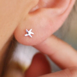 Può includere: Primo piano di un orecchino a forma di stella marina argentata su un orecchio. L'orecchino è piccolo e dettagliato, con cinque punte. Una mano con smalto rosso è in primo piano.