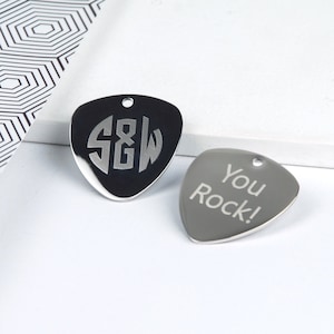 Puede incluir: Dos púas de guitarra plateadas con diseños grabados. Una púa tiene las iniciales "S&W" en un círculo. La otra púa tiene el texto "You Rock!" grabado.