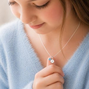 Peut inclure: Un collier en argent avec une chaîne fine. Le collier comporte un petit charme rond avec la lettre "A" et un pendentif en pierre précieuse bleue. La personne porte un pull bleu clair.