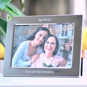 May include: A silver photo frame with a black mat and a photo of two women. The top of the frame has the text "Happy 70th Mum!" and the bottom has the text "You are, quite simply, the very best xxx".