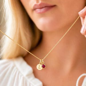 Op de afbeelding: Een goudkleurige ketting met twee bedels. Eén bedel is een kleine, ronde hanger met de letter "L" gegraveerd. De andere bedel is een kleine, ronde, rode edelsteen. De ketting wordt om de nek gedragen.
