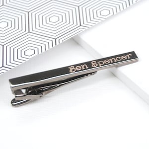 Personalised tie clip shown in gun metal finish with engraved name in modern curl font.