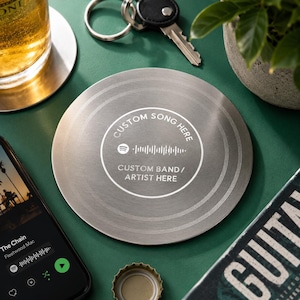 Op de afbeelding: Een ronde zilverkleurige metalen onderzetter met de tekst "CUSTOM SONG HERE" en "CUSTOM BAND / ARTIST HERE". De onderzetter heeft een Spotify-logo en een golfgrafiek. Een smartphone, een glas bier en sleutels zijn ook zichtbaar.