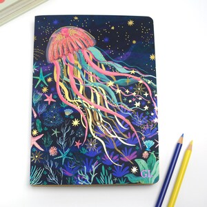 Può includere: Un taccuino con una vivace illustrazione di una medusa, stelle marine e piante acquatiche. La medusa è rosa e arancione con lunghi tentacoli fluenti in tonalità blu, gialle e oro. Lo sfondo è blu scuro con stelle e le iniziali "GL".