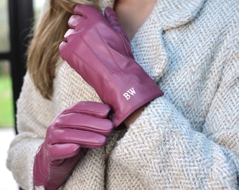 May include: Magenta leather gloves with the initials "BW" in white on the cuff. The gloves feature stitched detailing and are worn with a light tweed coat. The gloves are a deep pink colour.
